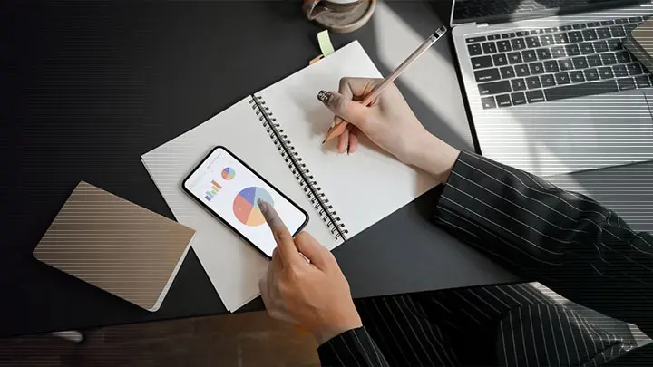 Eine Person macht sich Notizen in ein Notizbuch, daneben liegt ihr Handy mit einem Säulen- und Kreisdigramm.